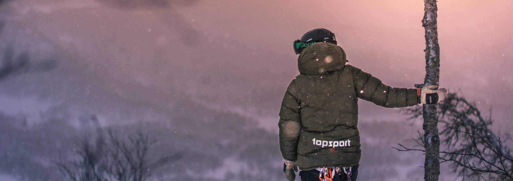 Person i vinterjacka med hjälm och handskar blickar ut över ett snötäckt fjällandskap i skymningsljus.